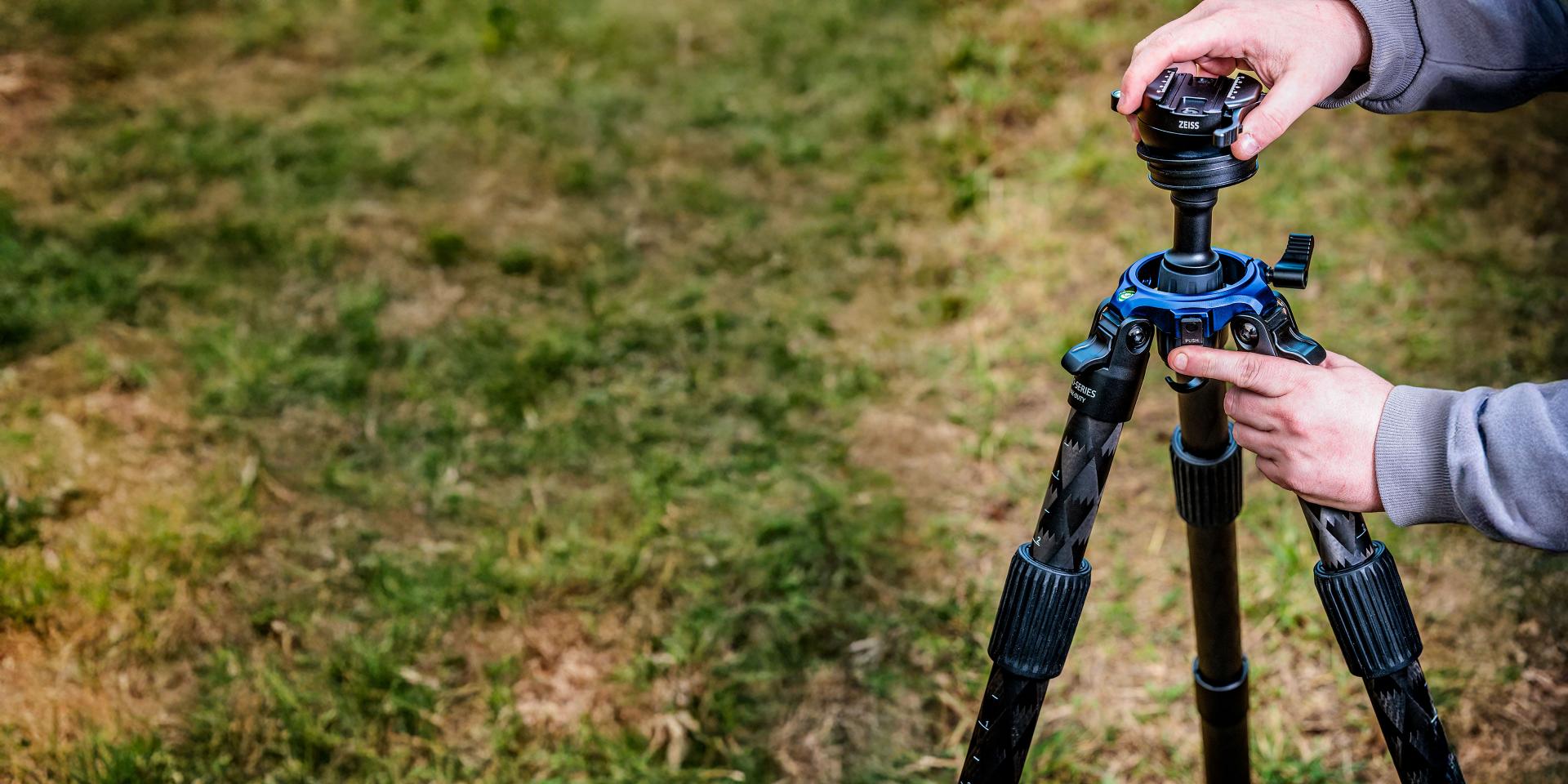 Person adjusting the head of a professional tripod setup on grass, with visible blue ZEISS components.