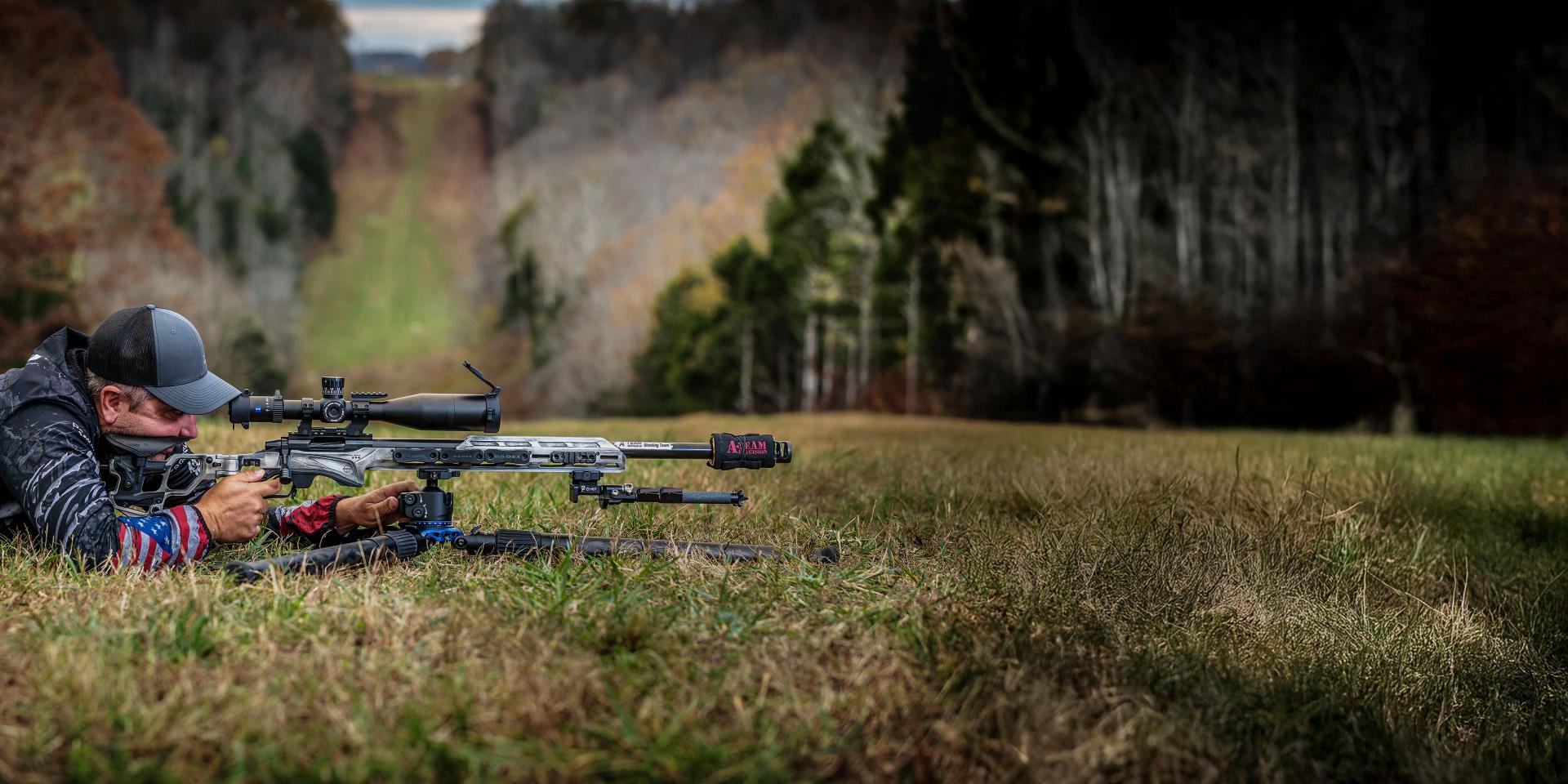 A man lies prone in a grassy field aiming a rifle equipped with a ZEISS scope, with a forested landscape in the background.