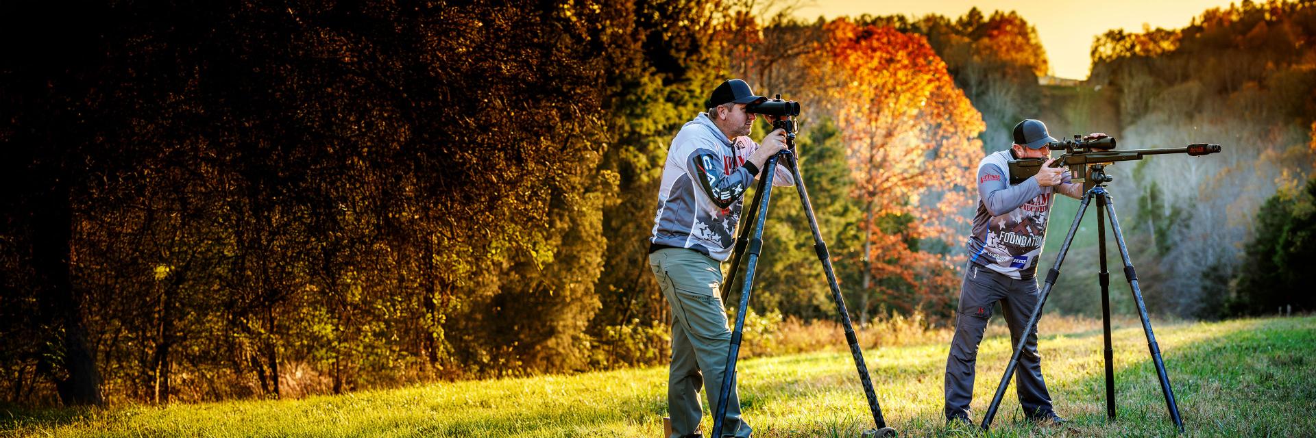 Two men are using ZEISS optical equipment mounted on tripods in a grassy field with a forest and autumn foliage in the background.