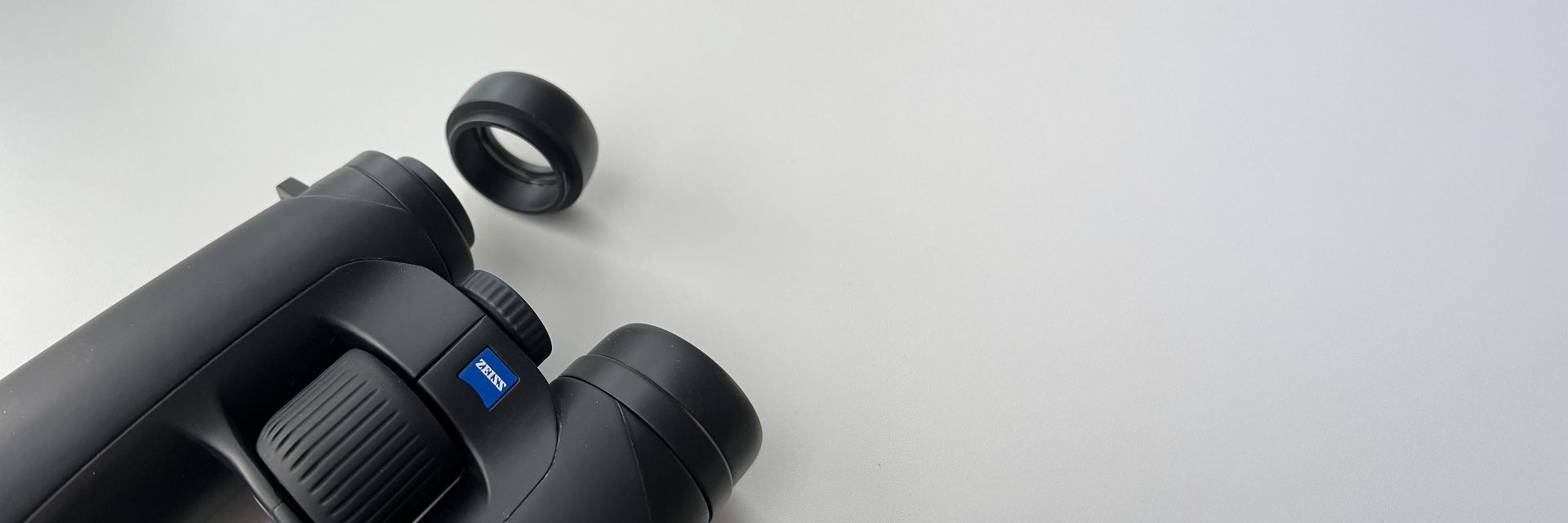 A pair of black binoculars featuring a textured grip and a detachable eyepiece placed nearby. The Zeiss logo is clearly visible on the binoculars, indicating the brand. The image is set against a clean, minimalistic white background, emphasizing the product's sleek design.