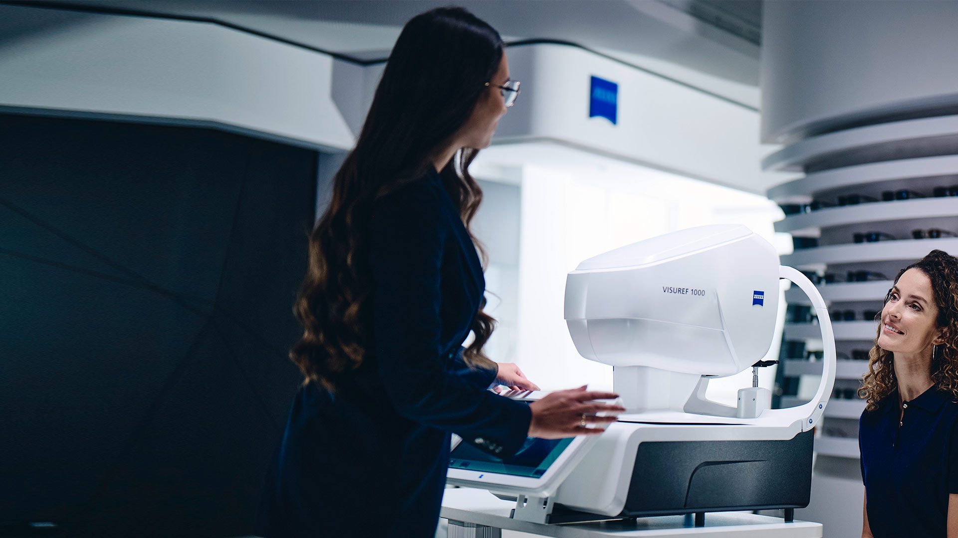 A woman sitting in front of the ZEISS VISUREF 1000 looking at a female optician.