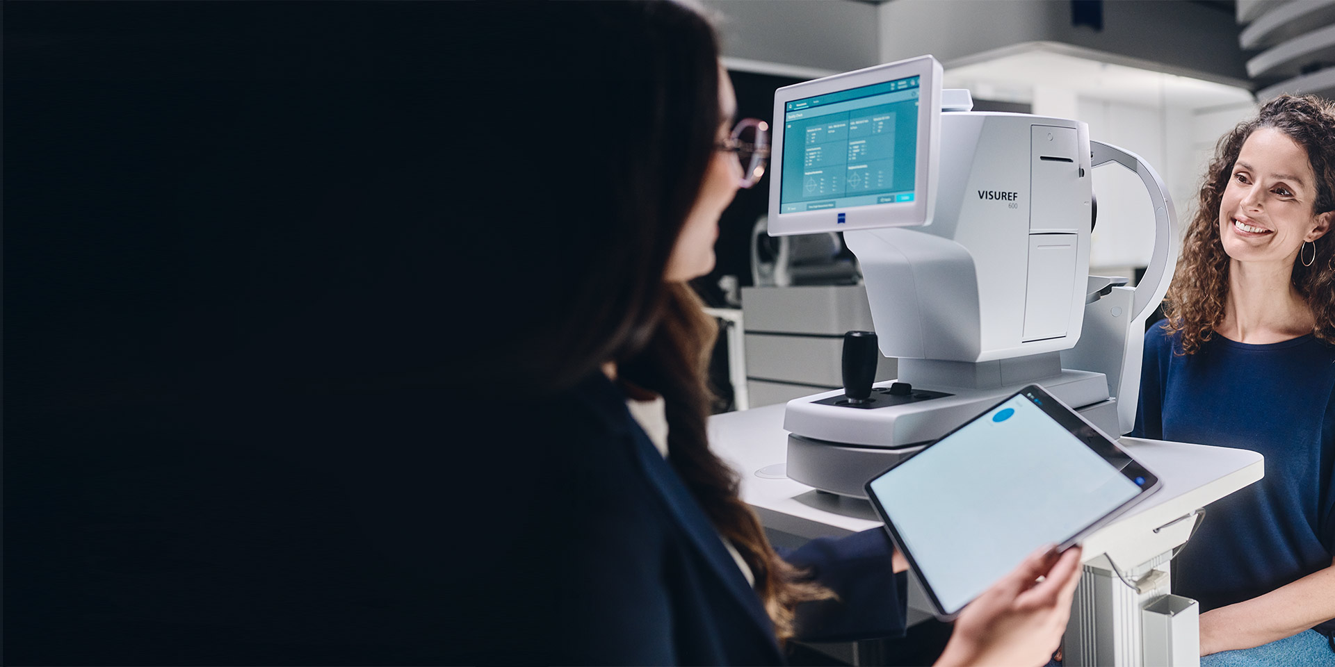 A female optician looking at her tablet sitting in front of the ZEISS VISUREF 600 and talking to her customer.