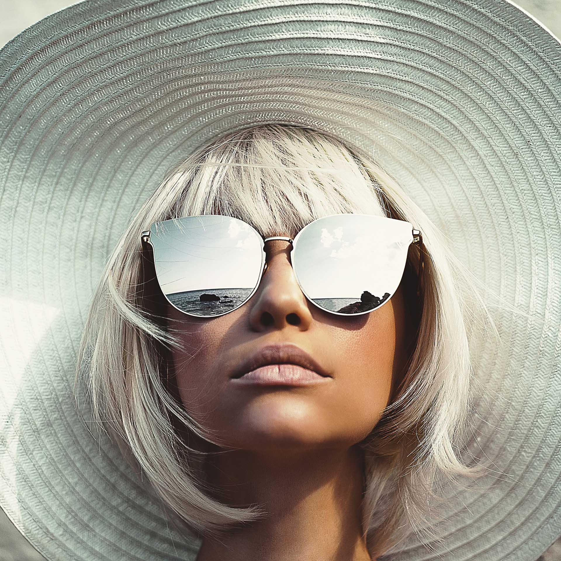 Young woman with big sun hat looks up and wears big sunglasses with mirror effect.​
